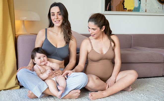 Zwei Mütter in Still-BHs sitzen auf dem Boden, eine davon schwanger, die andere hält ihr lachendes Baby auf dem Schoß